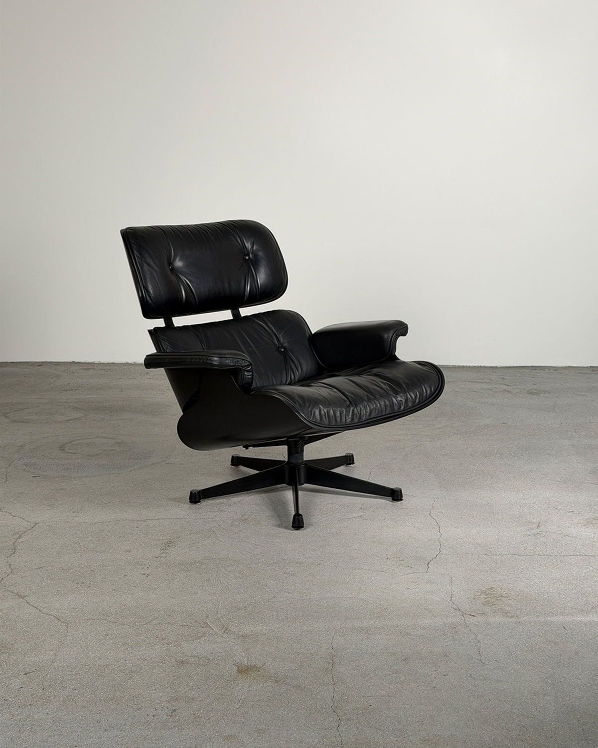 A classic black leather Eames-style lounge chair with a tufted headrest and five-star base, photographed in a minimalist studio setting with a concrete floor and plain wall.