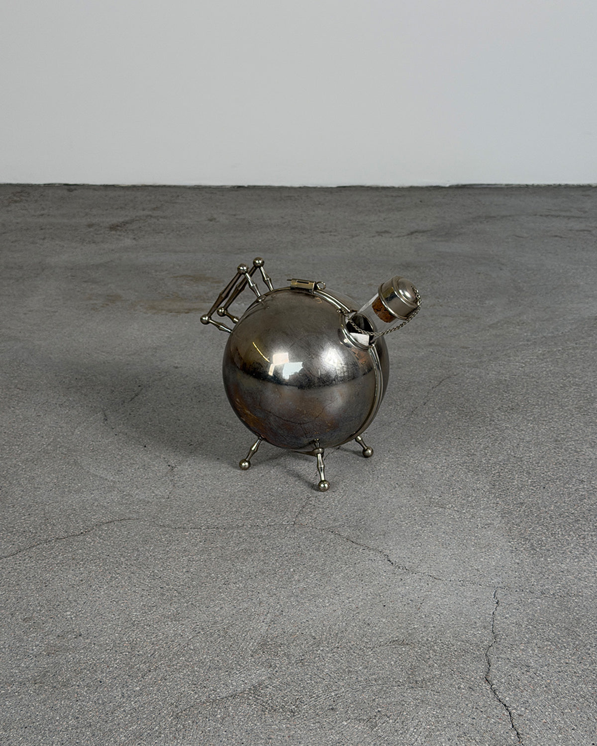 A polished silver metal decorative object with a rounded spherical body, small legs, and a curved spout, photographed on a gray concrete floor against a light gray wall.