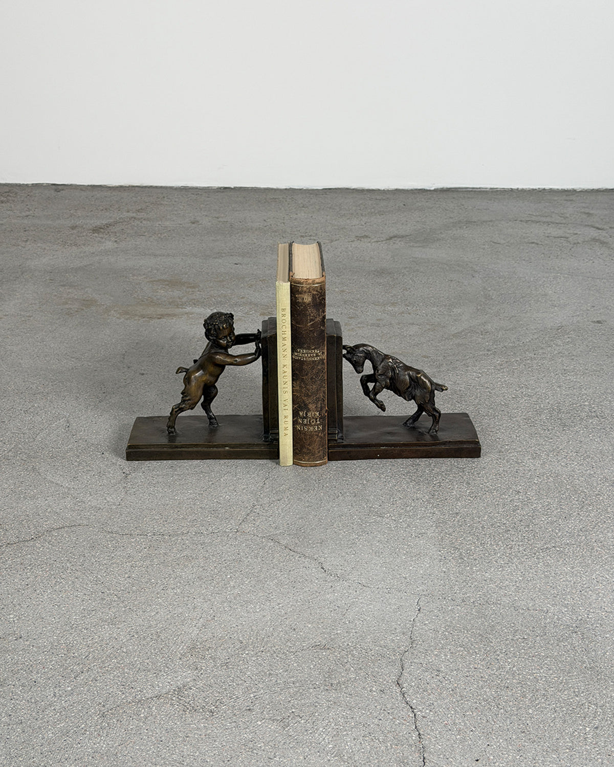 A pair of Vintage Bronze Bookends by Émile Joseph Carlier shaped like a satyr and a goat on a gray concrete floor against a white wall.