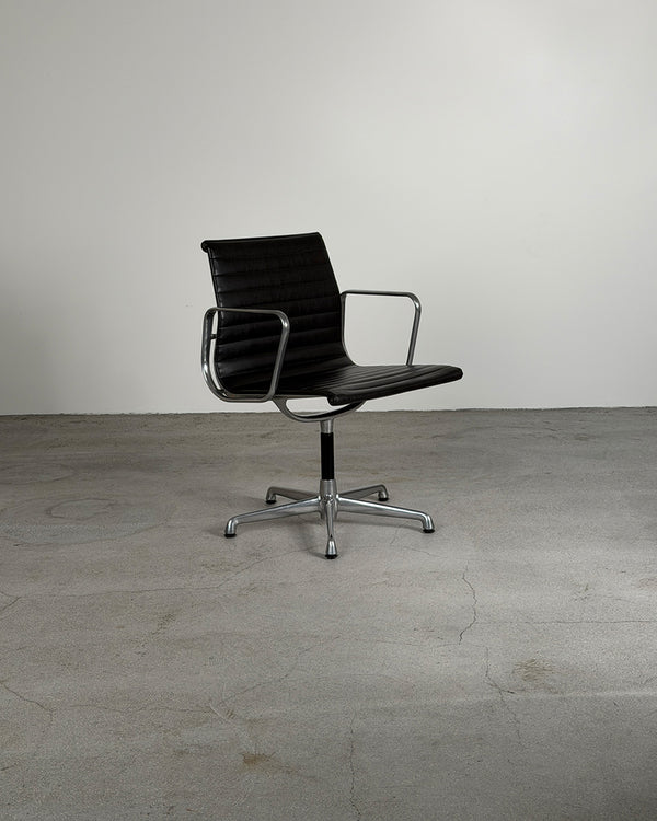 A modern black leather swivel office chair with tubular armrests and a chrome five-star base, set against a minimalist gray concrete floor and wall.