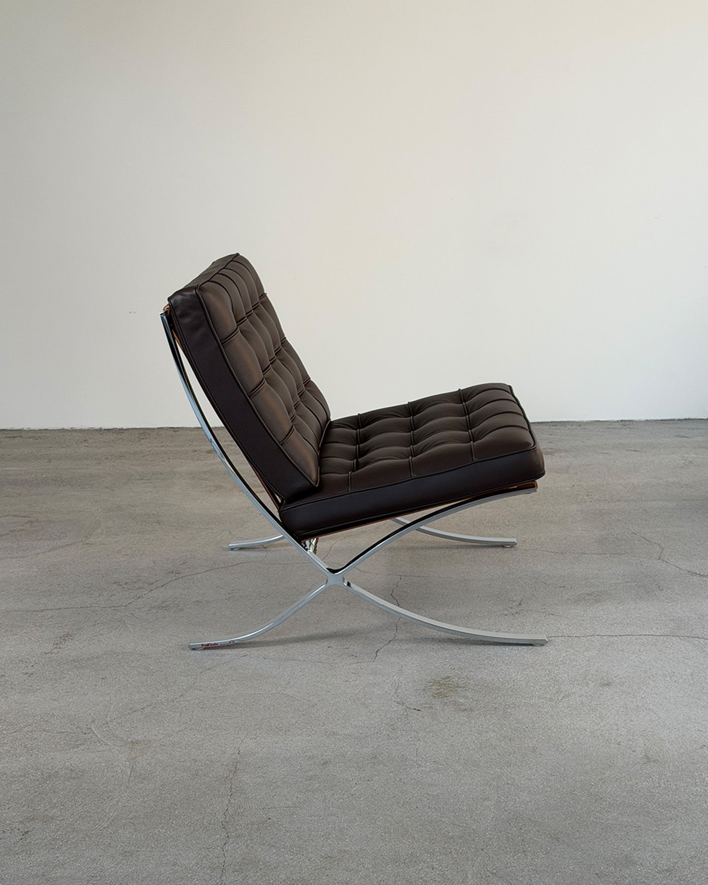 A modern Barcelona lounge chair with dark brown tufted leather upholstery and a chrome steel X-frame base, photographed in a minimalist studio setting with a concrete floor and plain off-white wall.