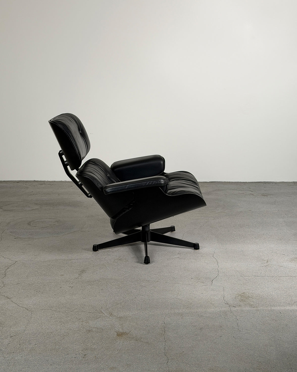A black leather lounge chair with a five-star swivel base, photographed in a minimalist studio setting with a concrete floor and a light gray wall.