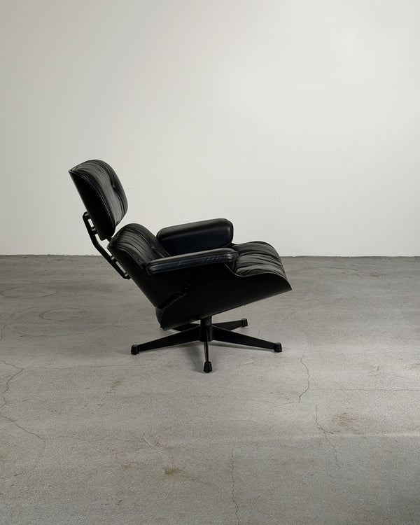 A black leather lounge chair with a five-star swivel base, photographed in a minimalist studio setting with a concrete floor and a light gray wall.