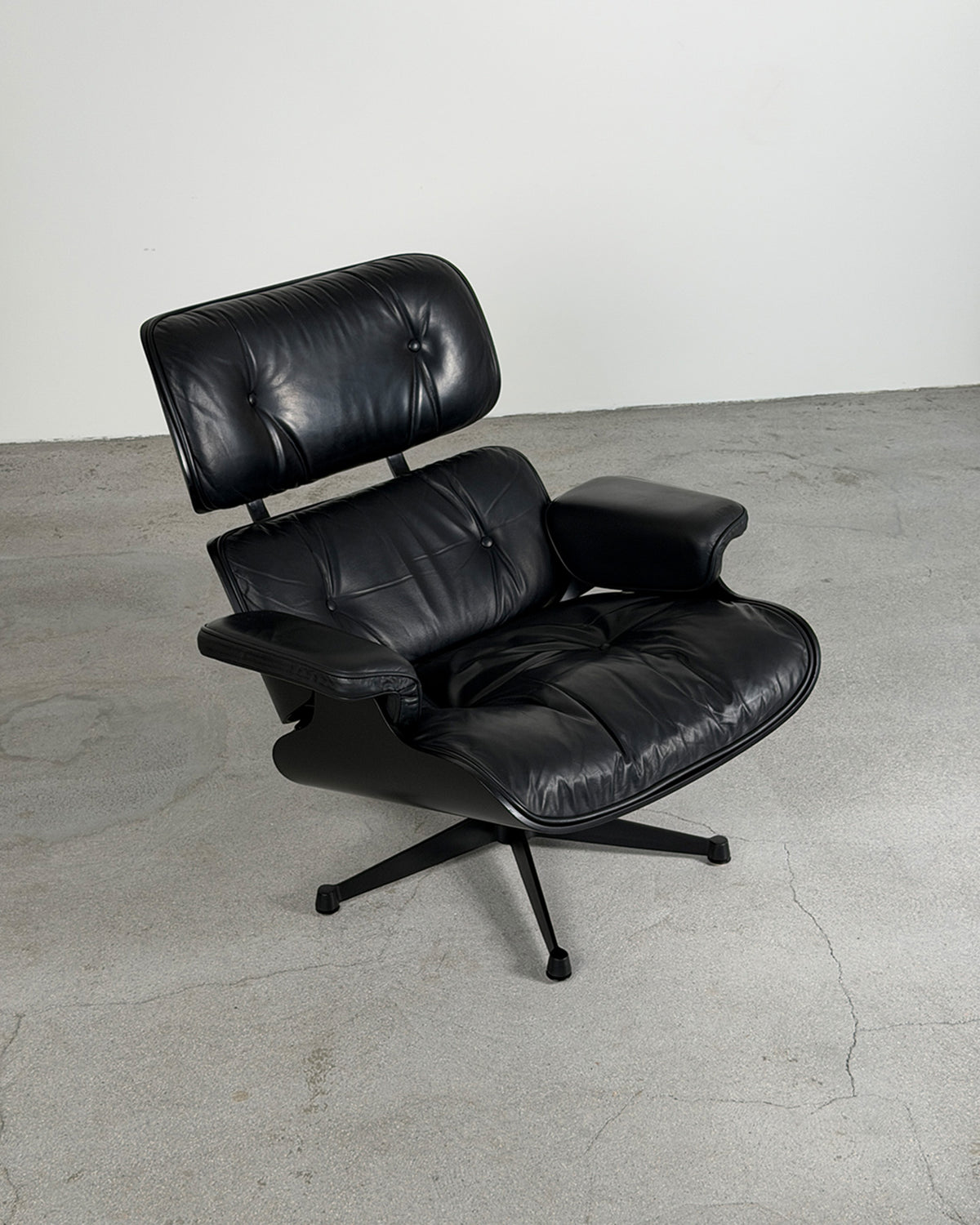 A Vitra Black Eames Lounge Chair by Charles & Ray Eames, 1980s with a tufted backrest and armrests, mounted on a black five-star swivel base, photographed in a minimalist studio setting with a concrete floor and white wall.