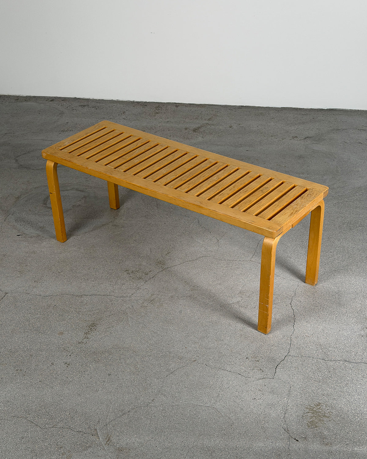 A rectangular wooden Artek bench with curved legs, photographed in a minimalist studio setting with a gray concrete floor and white wall.
