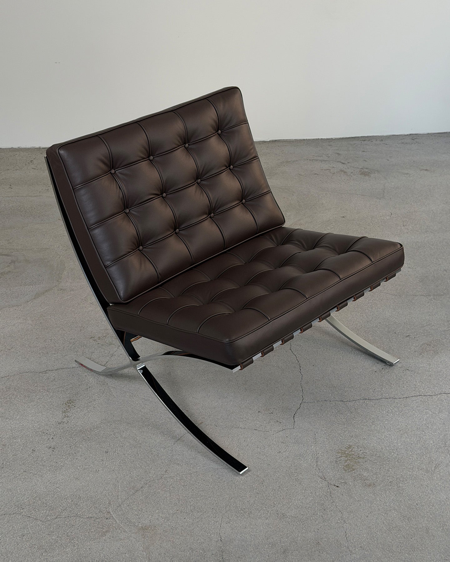 A modern brown leather armchair with a tufted backrest and seat, supported by a chrome S-shaped base, photographed in a minimalist studio setting with a concrete floor and plain wall.