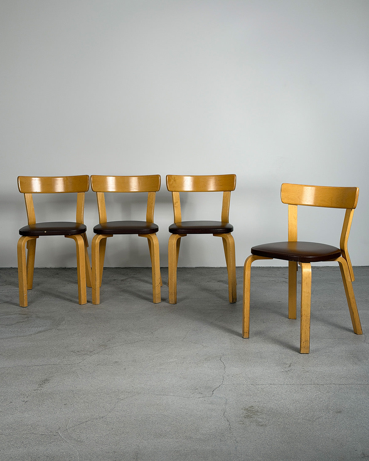 Four wooden chairs with brown seats on a gray floor and wall.