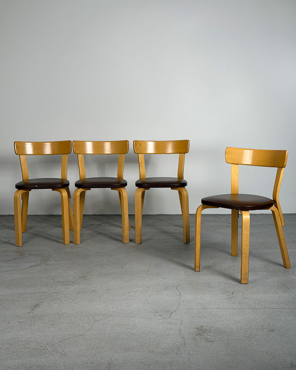 Four wooden chairs with brown seats on a gray floor and wall.