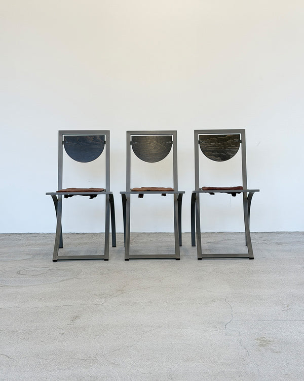 Three metal chairs with wooden seats on a light wooden floor against a white wall.