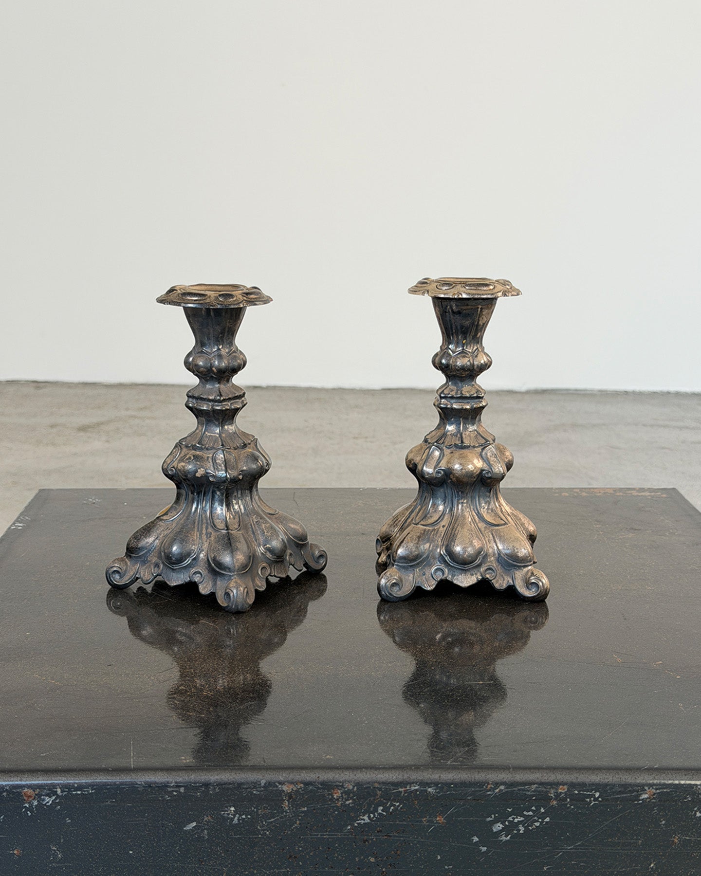 A pair of ornate antique-style metal candlesticks with sculpted floral bases, displayed side by side on a dark reflective surface against a plain light gray wall.