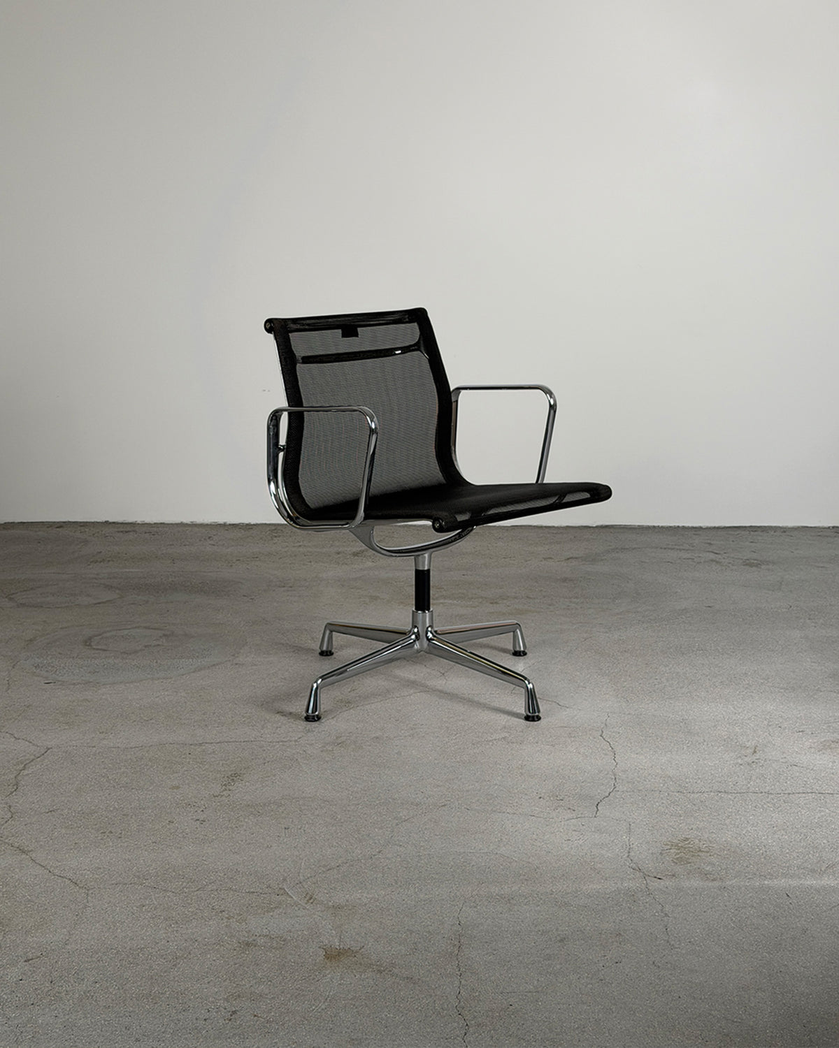 A modern Charles & Ray Eames EA108 Mesh Office Chair for Vitra with chrome armrests and a five-star metal base, photographed in a minimalist studio setting with a concrete floor and plain light gray wall.
