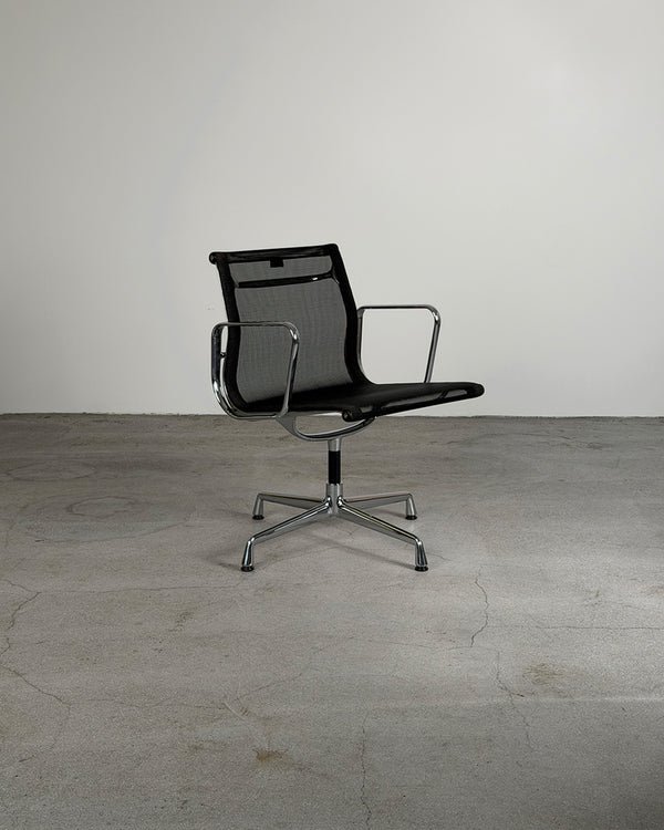A modern Charles & Ray Eames EA108 Mesh Office Chair for Vitra with chrome armrests and a five-star metal base, photographed in a minimalist studio setting with a concrete floor and plain light gray wall.