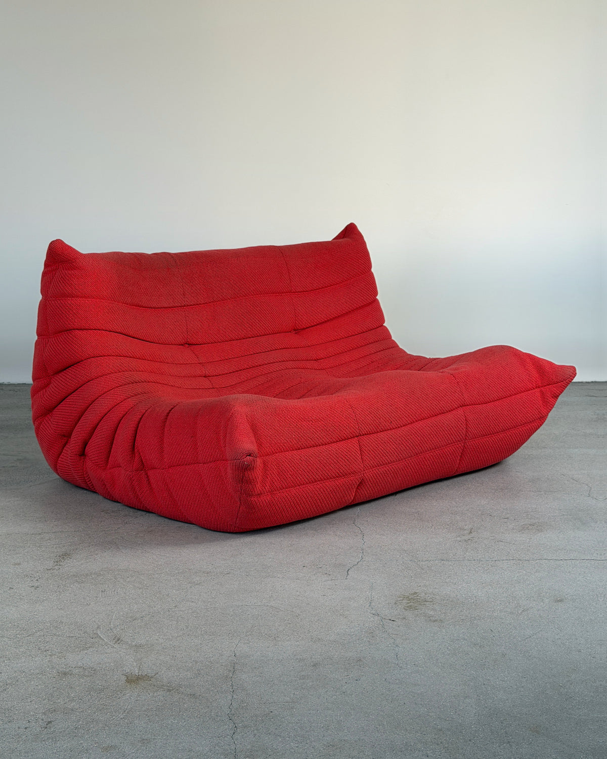 A bright red, low-profile lounge chair with a heavily padded, segmented tufted backrest and seat, set against a plain light gray wall and concrete floor.