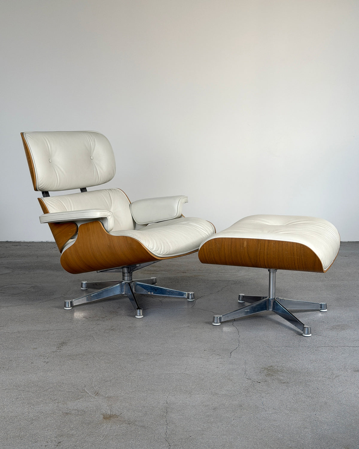 Mid-century modern Vitra Cream Eames Lounge Chair and Ottoman by Charles & Ray Eames, featuring cream leather upholstery and curved wood veneer shells on a metal swivel base, photographed in a minimalist studio setting.