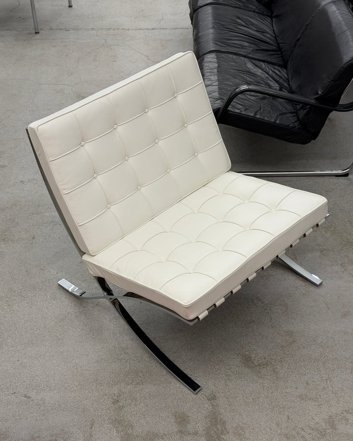 A modern Knoll Barcelona lounge chair with cream tufted leather upholstery and a curved chrome metal base, photographed on a concrete floor next to a black leather sofa.